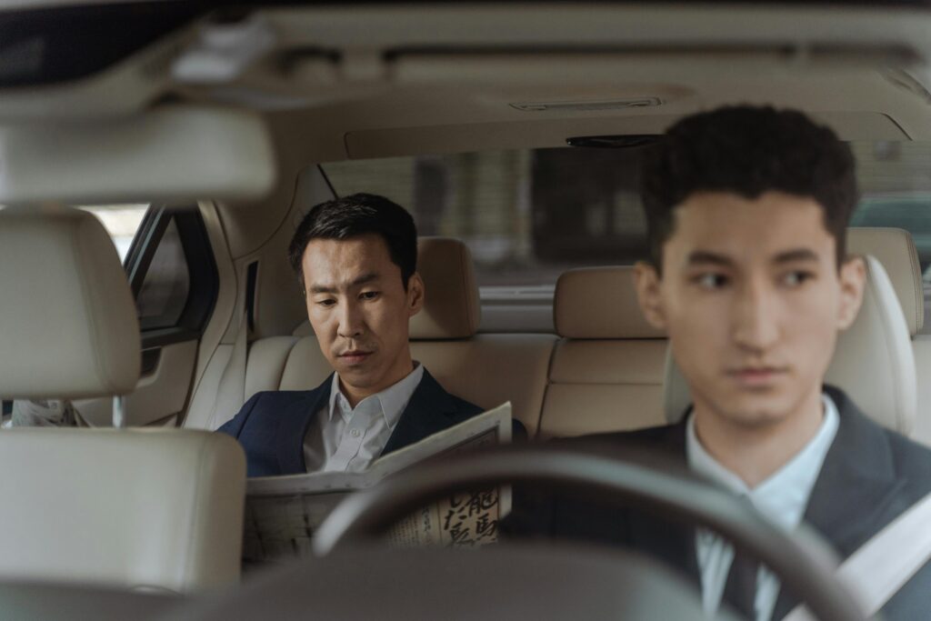 Elegant Asian businessman reading a newspaper inside a luxury car. Professional driver in focus.