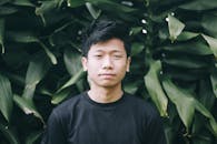 A young man stands in front of lush green foliage wearing a black t-shirt.